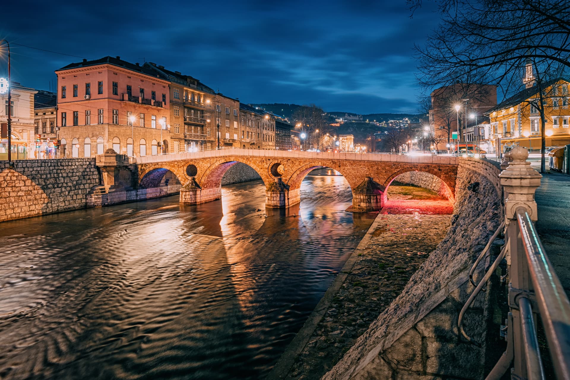 The Latin Bridge over the Miljacka River in Sarajevo at dusk, its stone arches reflected in the water