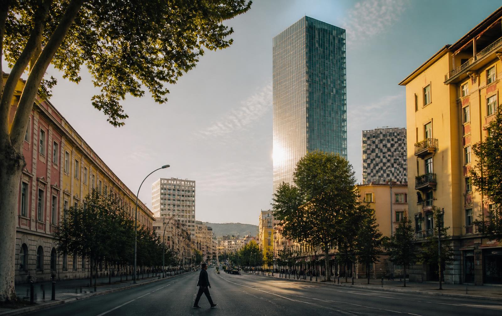 A boulevard in Tirana in late afternoon light, a single figure crossing
